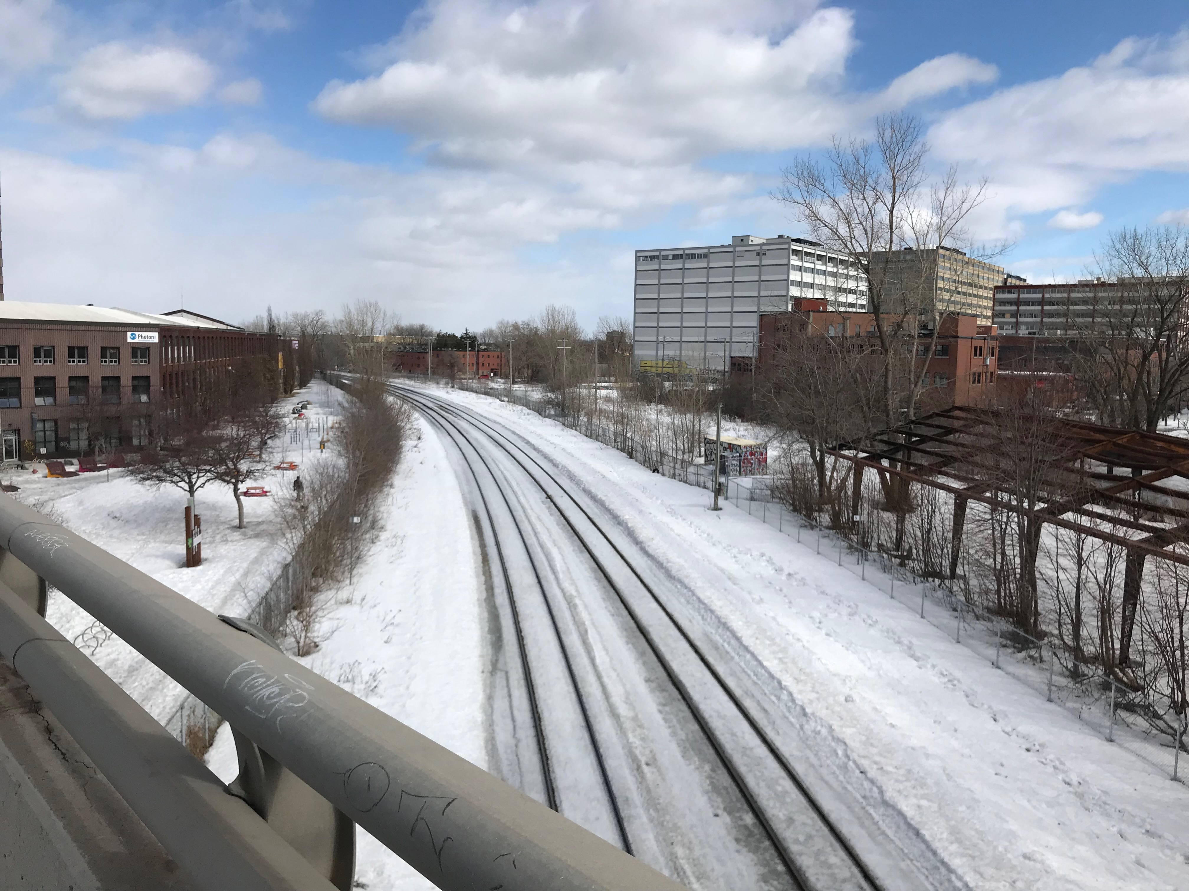 a photo of some snowy traintracks, a blue cloudy ski, and buildings.